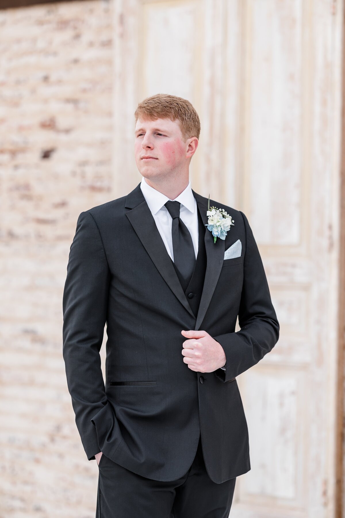 A groom in his black suit stands with his hand in his pocket and jacket coat off to the side over his shoulder.