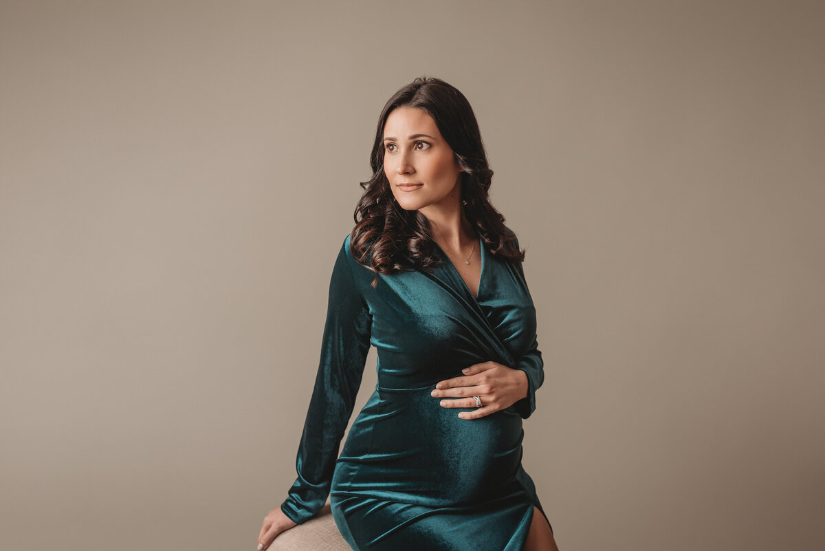 Pregnancy portrait of a woman wearing velvet green maternity gown sitting on stool looking over shoulder