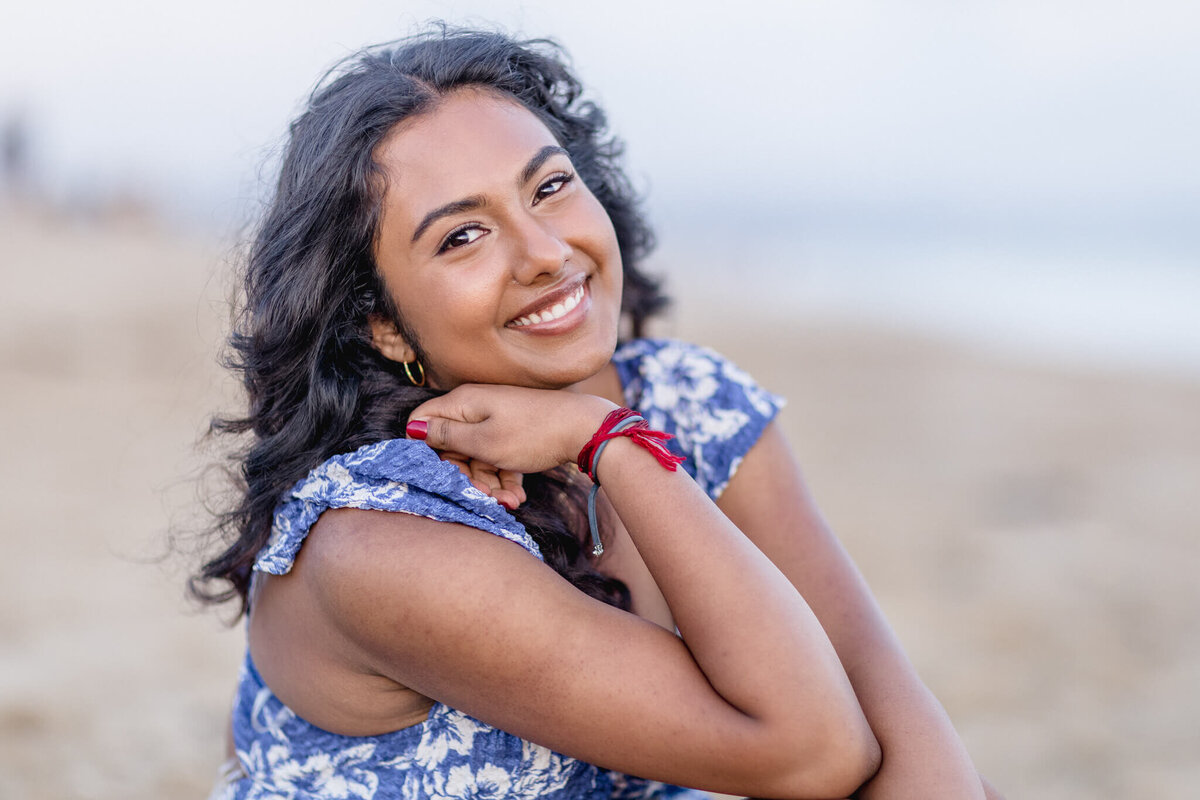A woman with long curly hair and a blue floral dress smiles at the camera. She rests her chin on her crossed arms, sitting on a sandy beach with the ocean in the background.