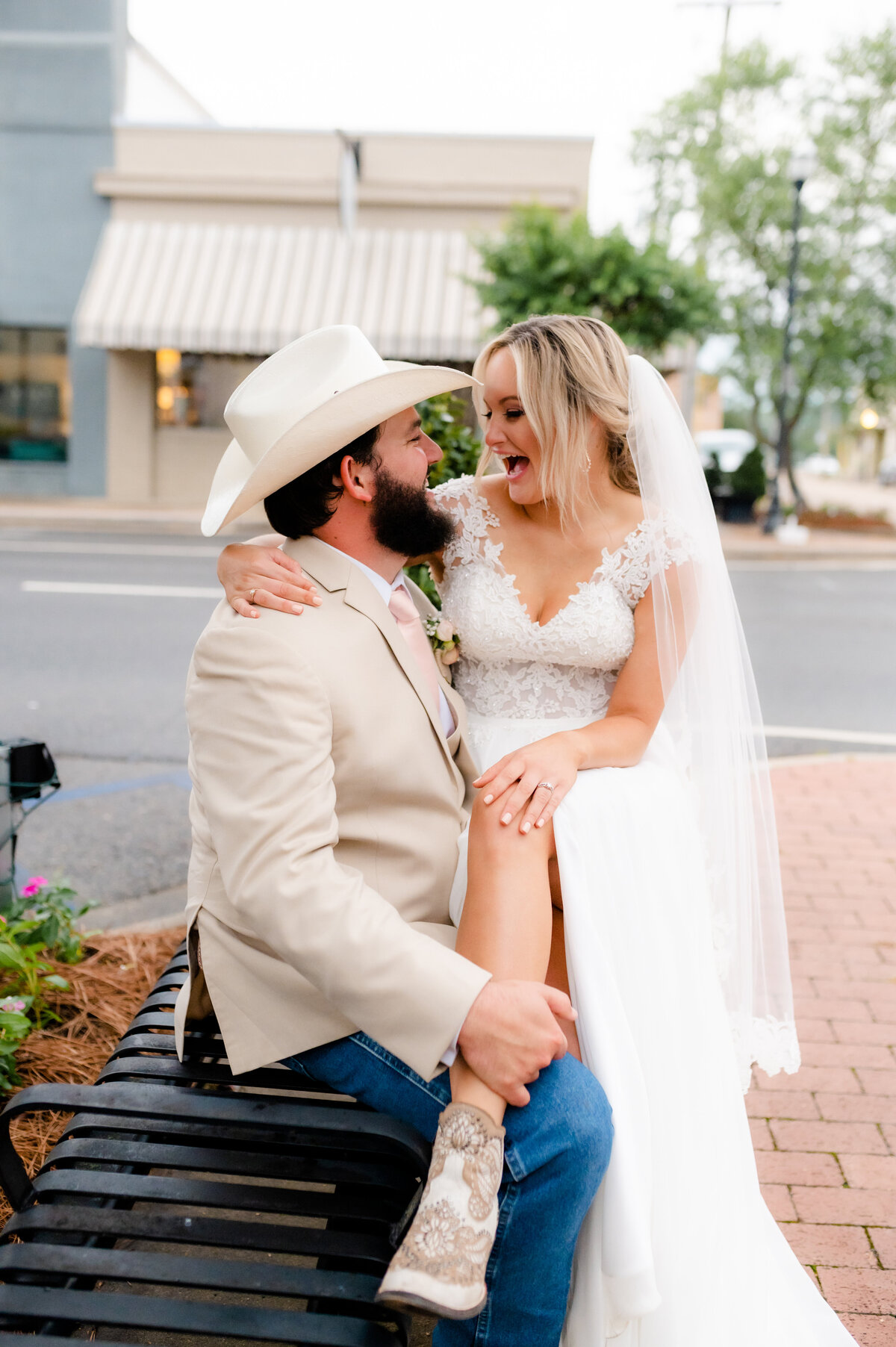 grooms sits on a bench downtown while his bride sits on his lap and laughs with him