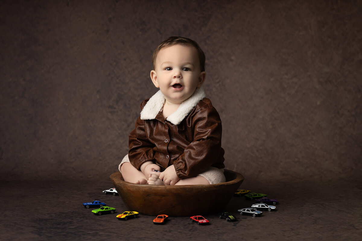 One-year-old boy wearing a brown aviator jacket and sitting in a wooden bowl, surrounded by colorful toy cars on a brown backdrop, captured by a Dallas milestone photographer.