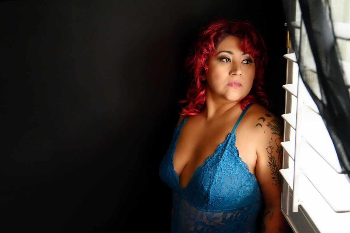 Red-haired woman wearing a teal blue lingerie staring out the window.