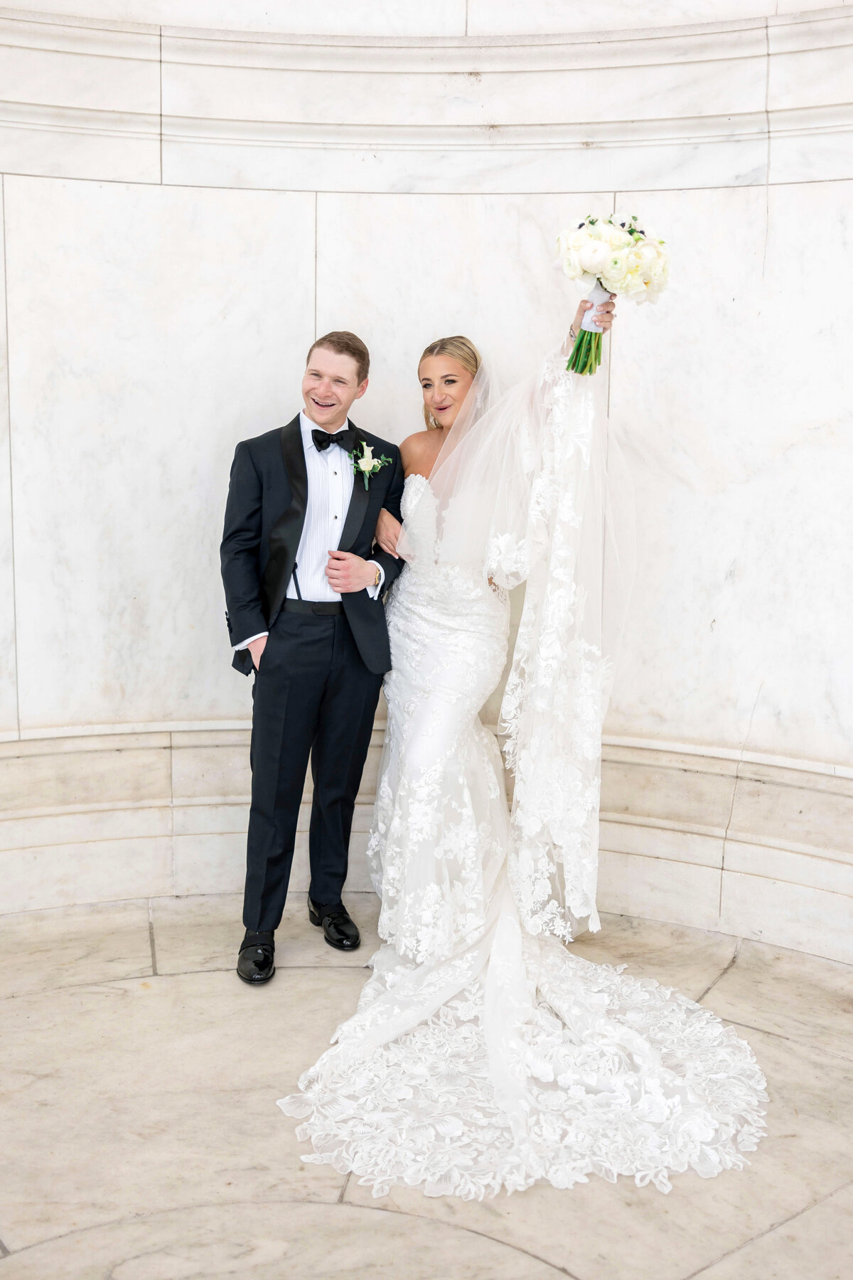 A bride and groom pose in formal attire against a marble backdrop. The bride, in a flowing lace gown and veil, holds a bouquet aloft while smiling. The groom wears a black tuxedo and bow tie, with an arm linked with the brides.