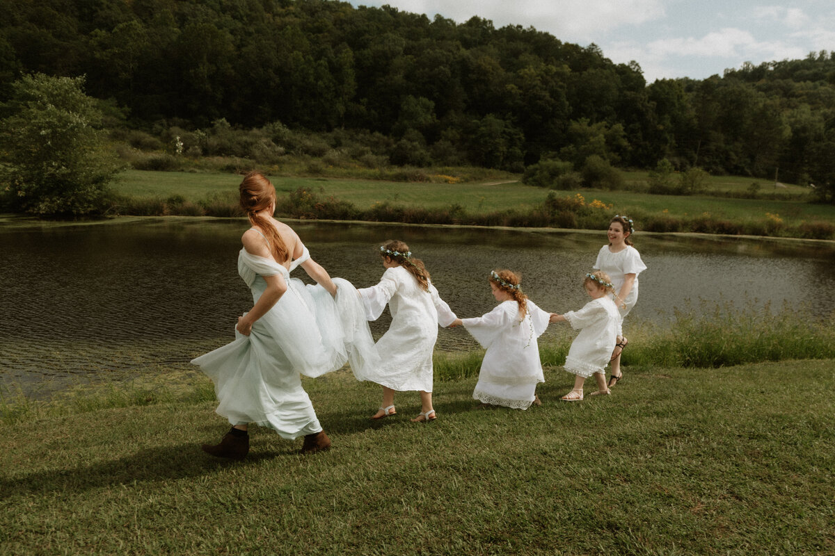Flower girls and bridesmaids in handmade dresses running hand in hand beside a lake during a West Virginia mountain wedding, captured by Rainstorm Photo & Video with fairytale, Pride and Prejudice-inspired charm.
