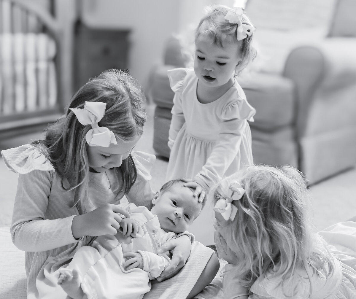 Newborn lifestyle photography, black and white sibling portrait, Birmingham newborn photographer – A heartwarming black and white image of three older sisters joyfully surrounding their newborn sibling on a cozy bed, capturing the essence of family love and connection.