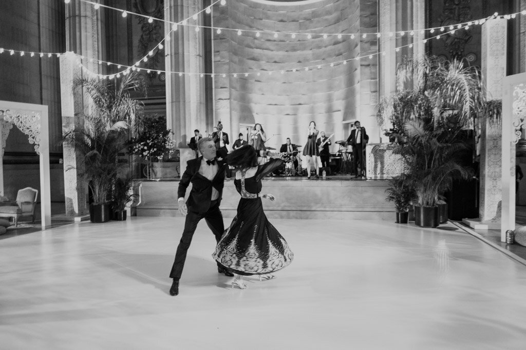 This black-and-white photo of a couple dancing on an elegantly decorated dance floor captures a moment at a formal event at Washington D.C.'s The Mellon Auditorium, one of the best luxury DC venues with European flair. The man is dressed in a stylish suit, while the woman is wearing a flowing dress, creating a classic and sophisticated look. In the background, a live band can be seen performing on a stage, adding to the ambiance of the evening. Beautiful string lights hang across the venue, casting a warm and romantic glow over the scene, beautifully captured by photographer Sarah Bradshaw.