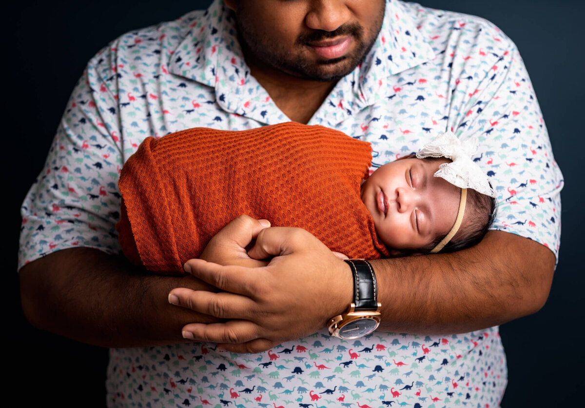 A newborn baby girl wrapped in orange sleeps in her daddys arms