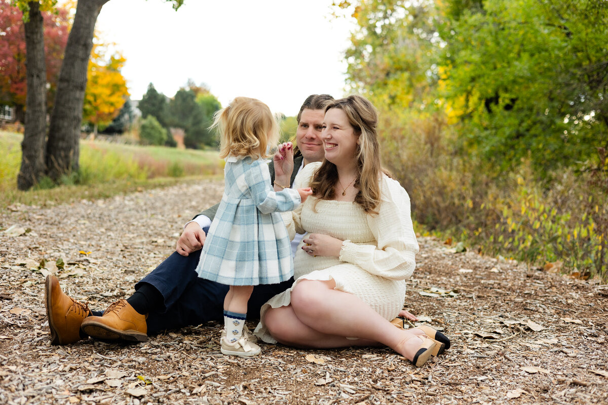 Mom and dad sit on the ground and smile at toddler daughter while feeding her a snack.