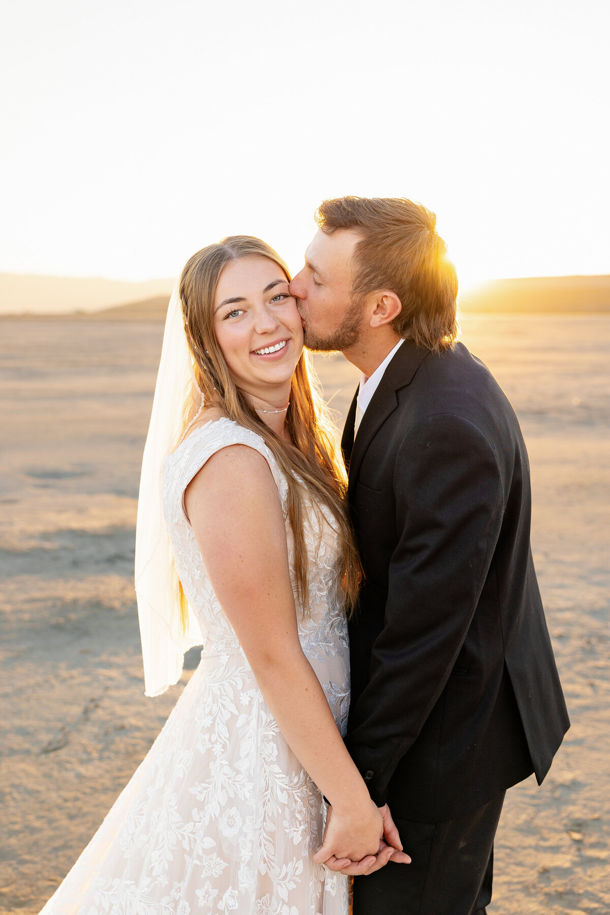 Groom kisses the bride’s cheek as she smiles at the camera, bathed in warm morning light.