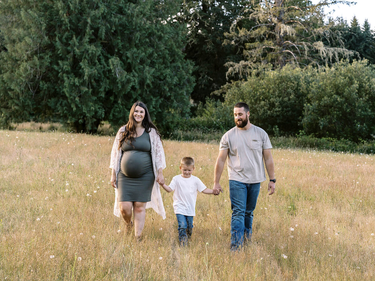 A pregnant woman, her son and husband hold hands in a field during a maternity photography shoot.
