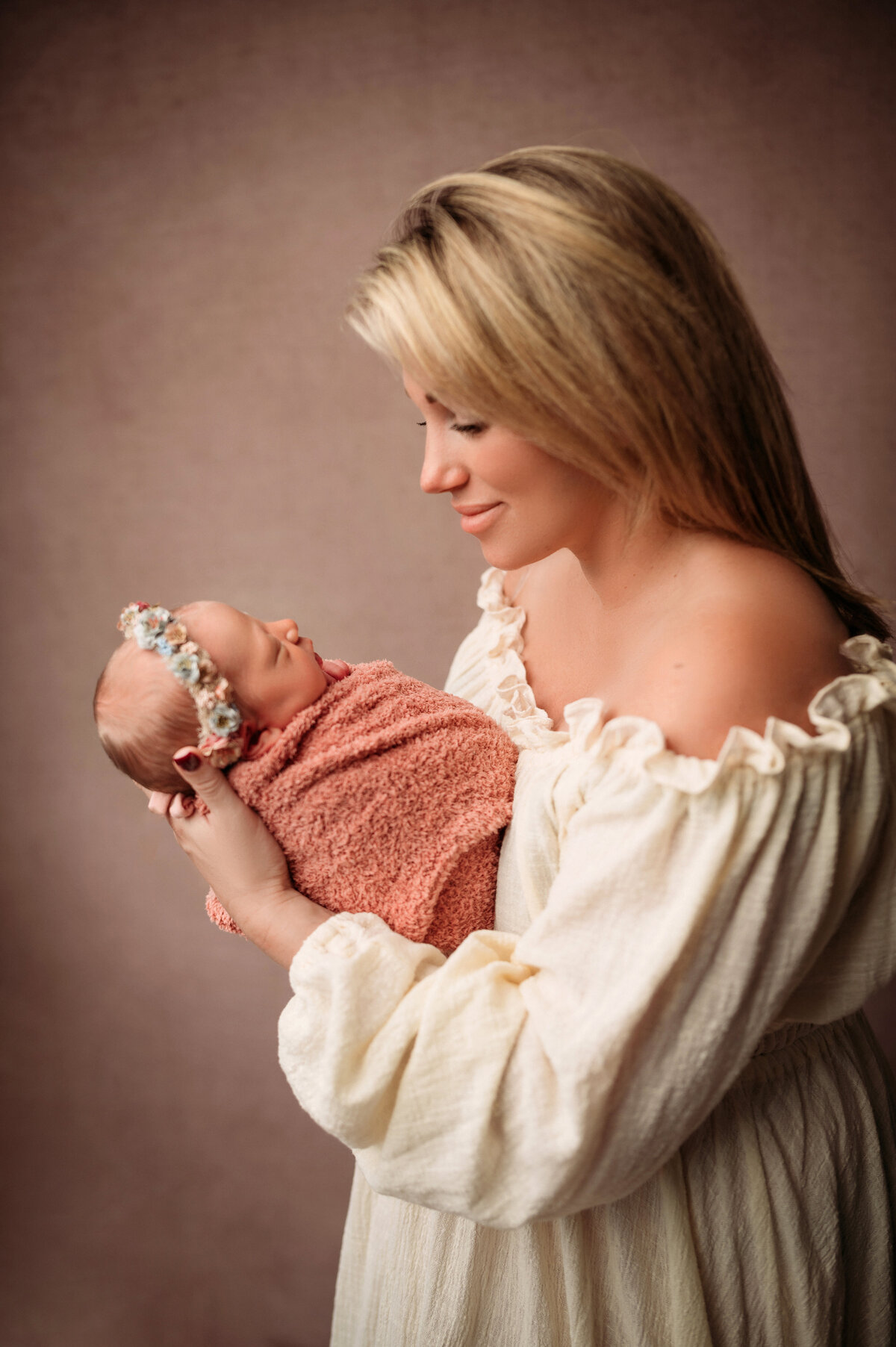 new mom with short blonde hair wearing a cream peasant gown holding swaddled newborn baby girl in a pink swaddle and floral headband looking at mom on brown backdrop