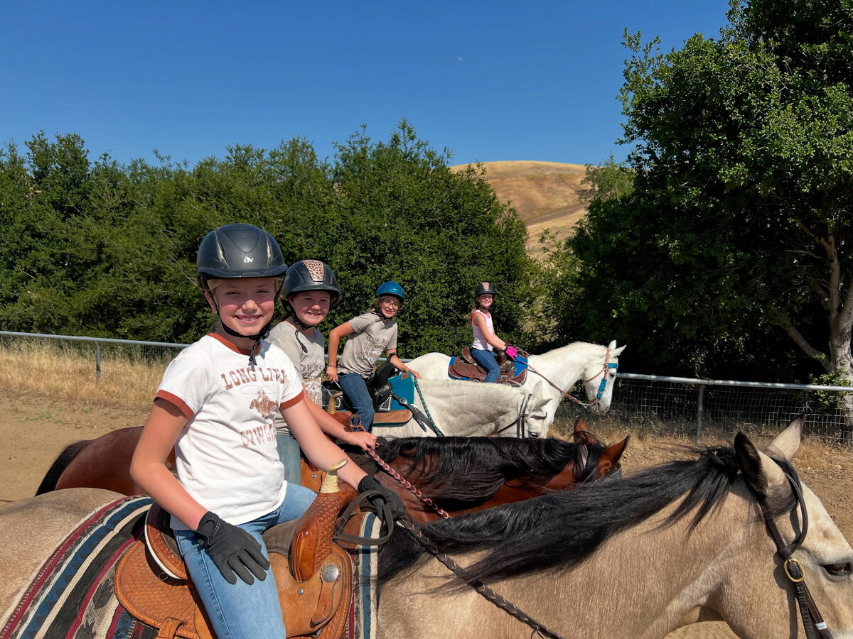 A group of four young riders, each sitting on a horse, they are riding in western saddles and smiling for the camera