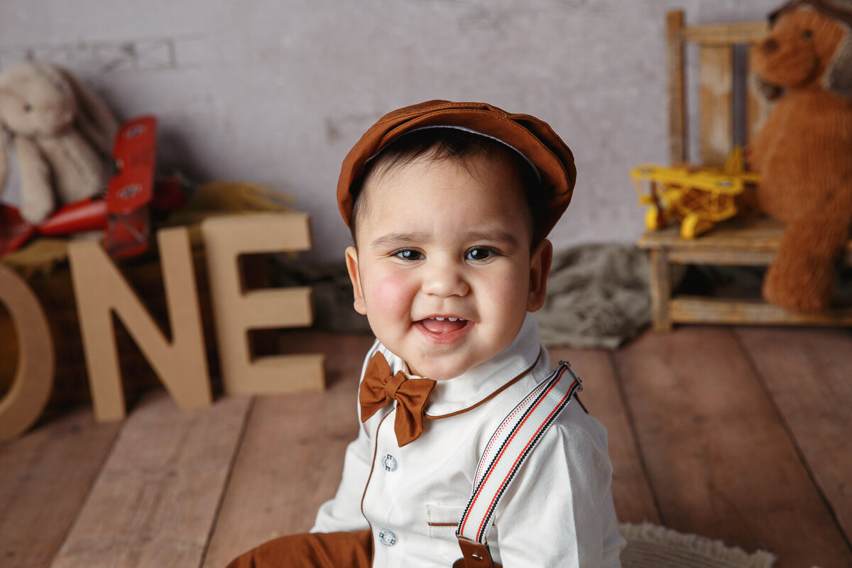 Close up of a smiling one year old wearing a cut little cap and suspenders photographed on a background with teddy bears