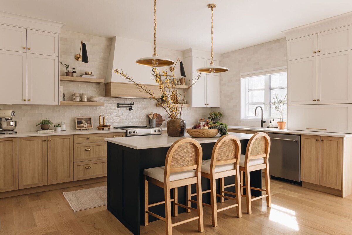 Bright modern kitchen in Montreal featuring light wood lower cabinets, white upper cabinetry, black island, and brass pendant lights – by Ingrid Interior Design.