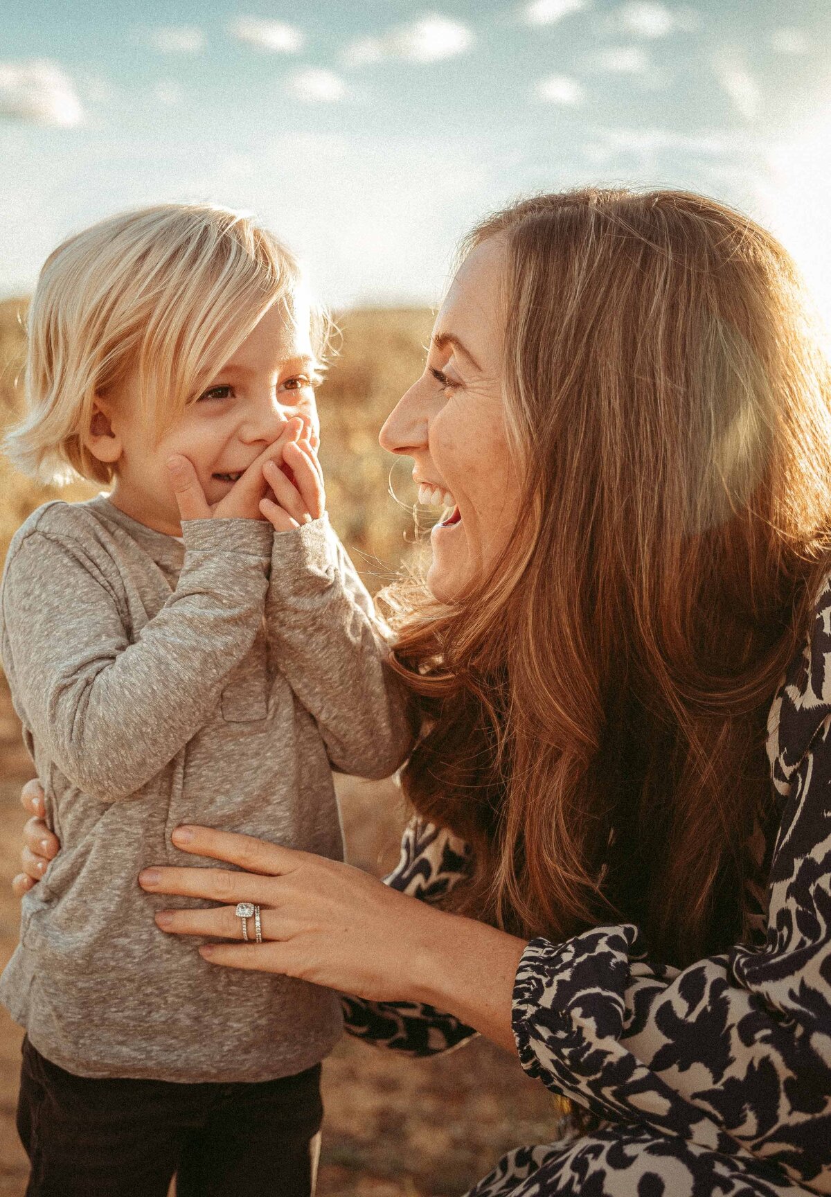 Young boy is laughing and covering his mouth with both of his hands while his mother laughs and looks at him
