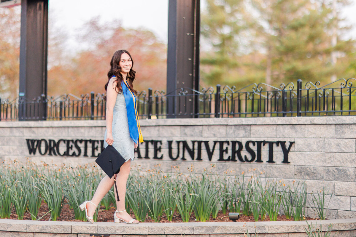 A girl holding a graduation cap and walking in front of her college's sign representing Boston cap and gown photography