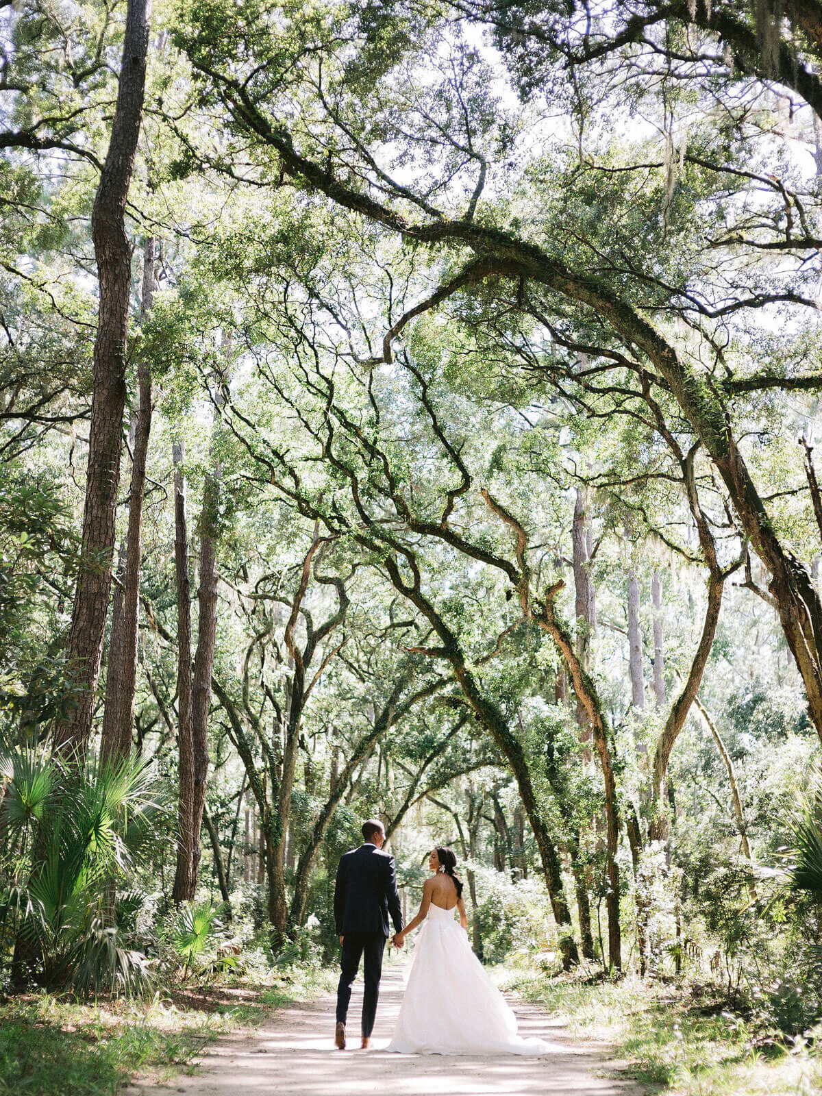 The bride and groom are walking hand in hand amongst the trees in Montage at Palmetto Bluff. Destination wedding image by Jenny Fu Studio