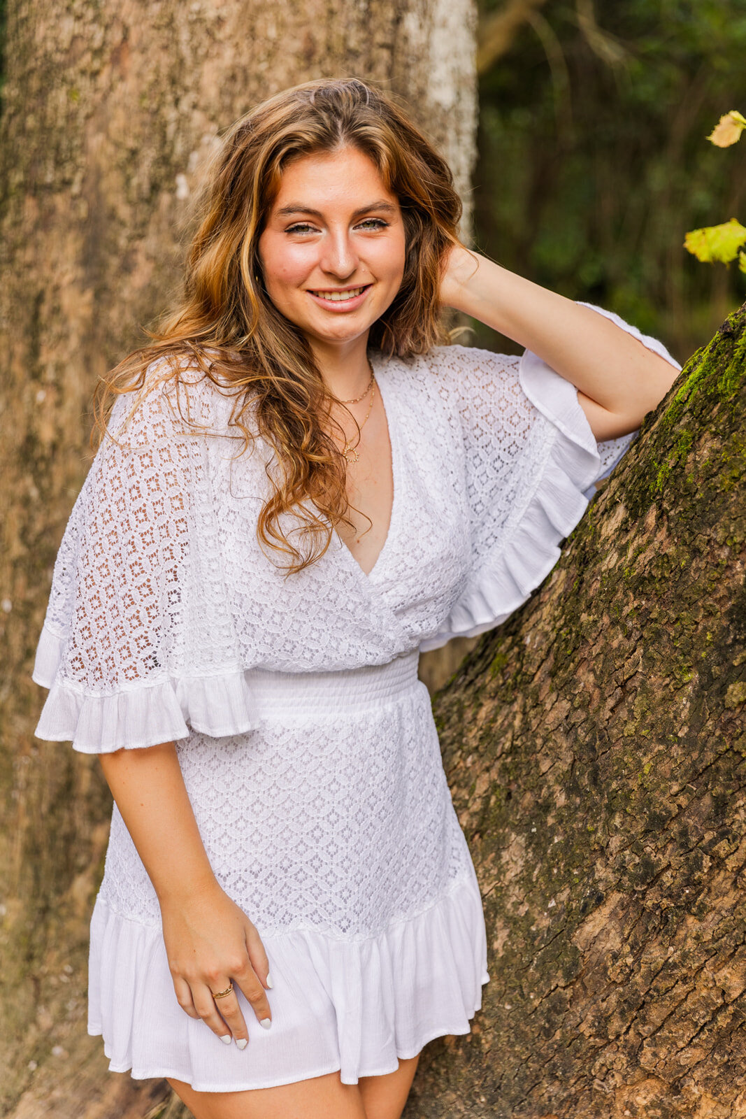 High shcool girl leaning against a tree, hand in her hair wearing a white dress during summer photo session by Laure Photography