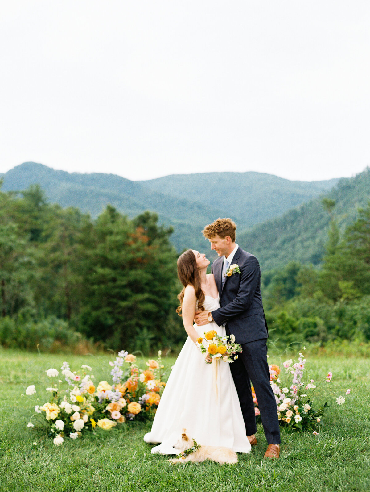 The bride and groom gaze at each other after their wedding, with their dog in front of the dress and the wedding arrangements behind, framed amongst the hills of North Carolina as seen from Paint Rock Farm, on medium format film by destination photographer My Sun and Stars Co.