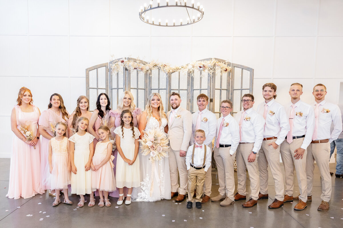The bride and groom stand in the center, surrounded by bridesmaids in soft pink and groomsmen in beige and white, all smiling at the camera.