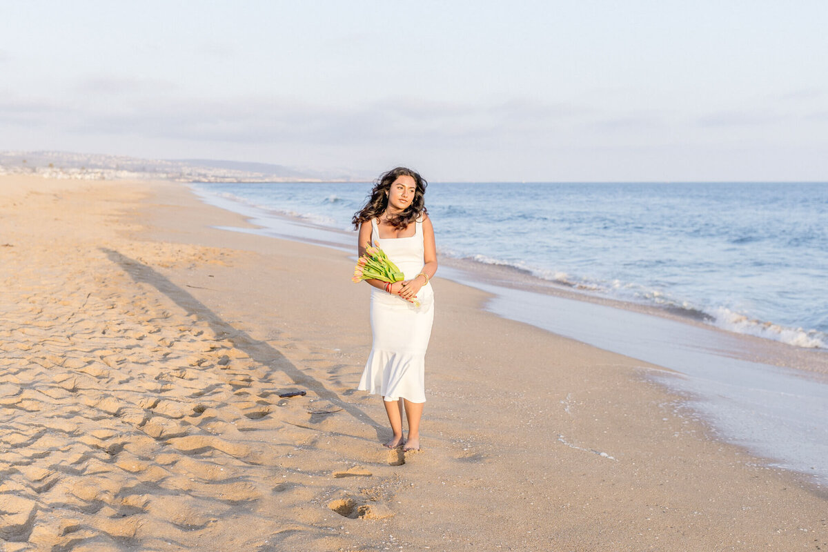 A woman in a white dress walks barefoot on a sandy beach, holding a bouquet of yellow flowers. The ocean waves gently lap at the shore, and the sky is clear with a few clouds.
