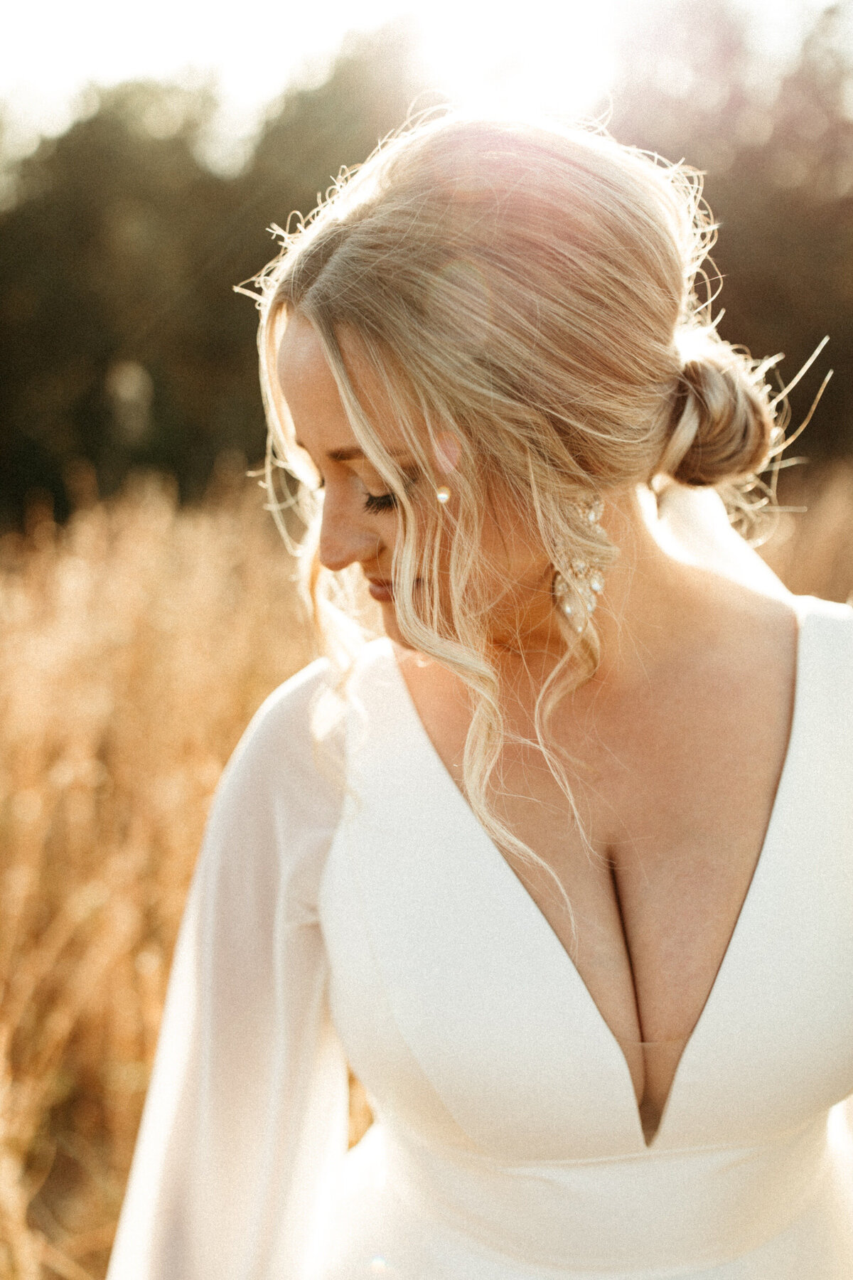 Elegant bride with messy updo and a wedding dress with long sleeves looking down over her shoulder in a field with the sun behind her