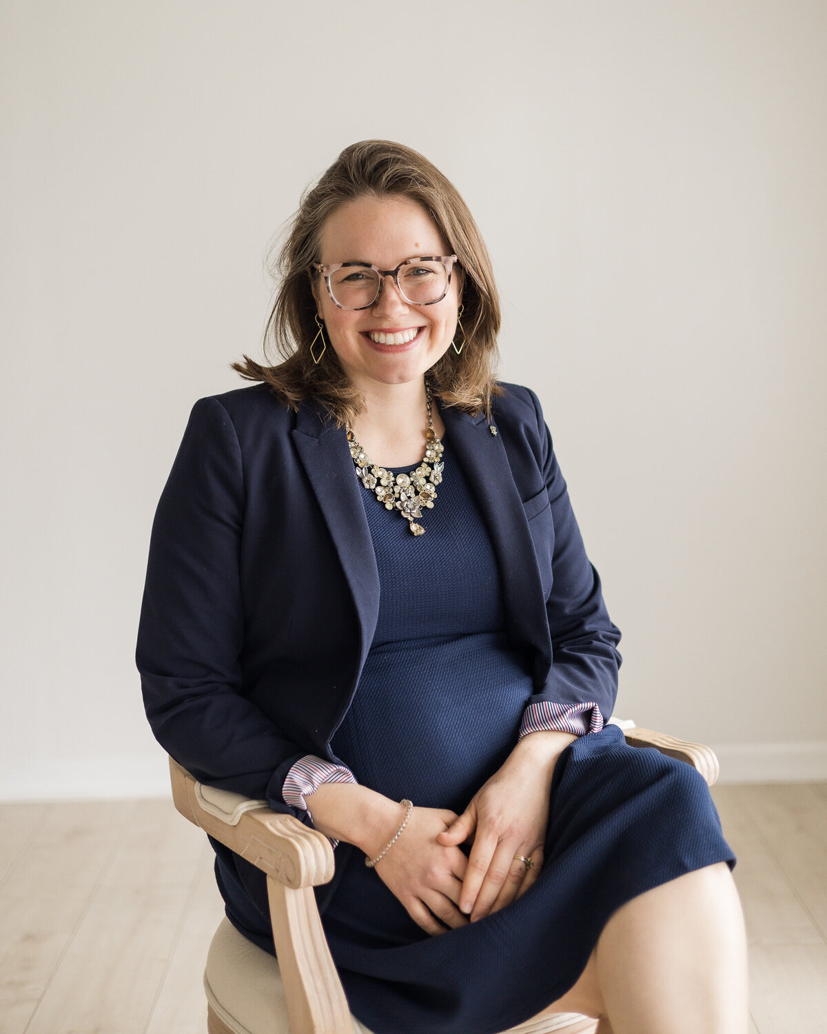 Brunette blonde pregnant female sitting in a chair, wearing glasses, a dark blue dress with a long sleeve dark blue jacket, smiling at the camera