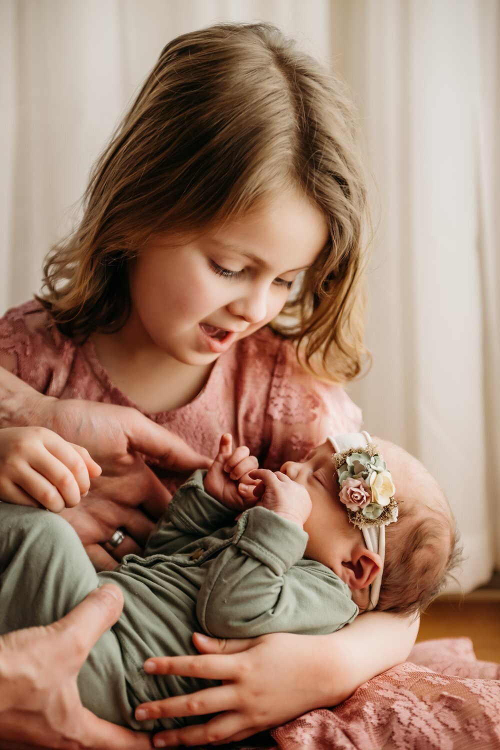 mom hands a toddler her baby sister. sister looks happily down at the baby