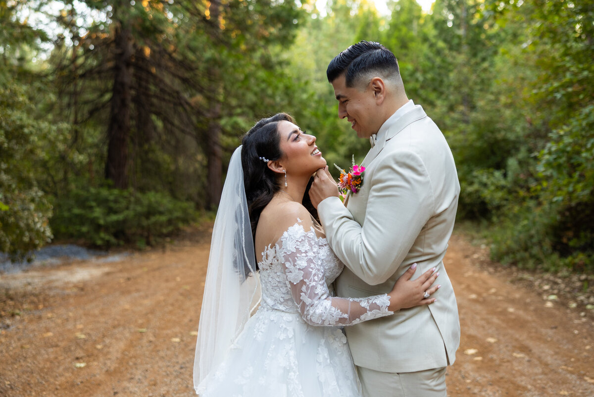 Bride and groom stand on a path near forest house lodge with forest trees in the background and look at each other while holding each other. The groom places a hand under the bride's chin. Photo by wedding photographer sacramento, CA philippe studio pro.