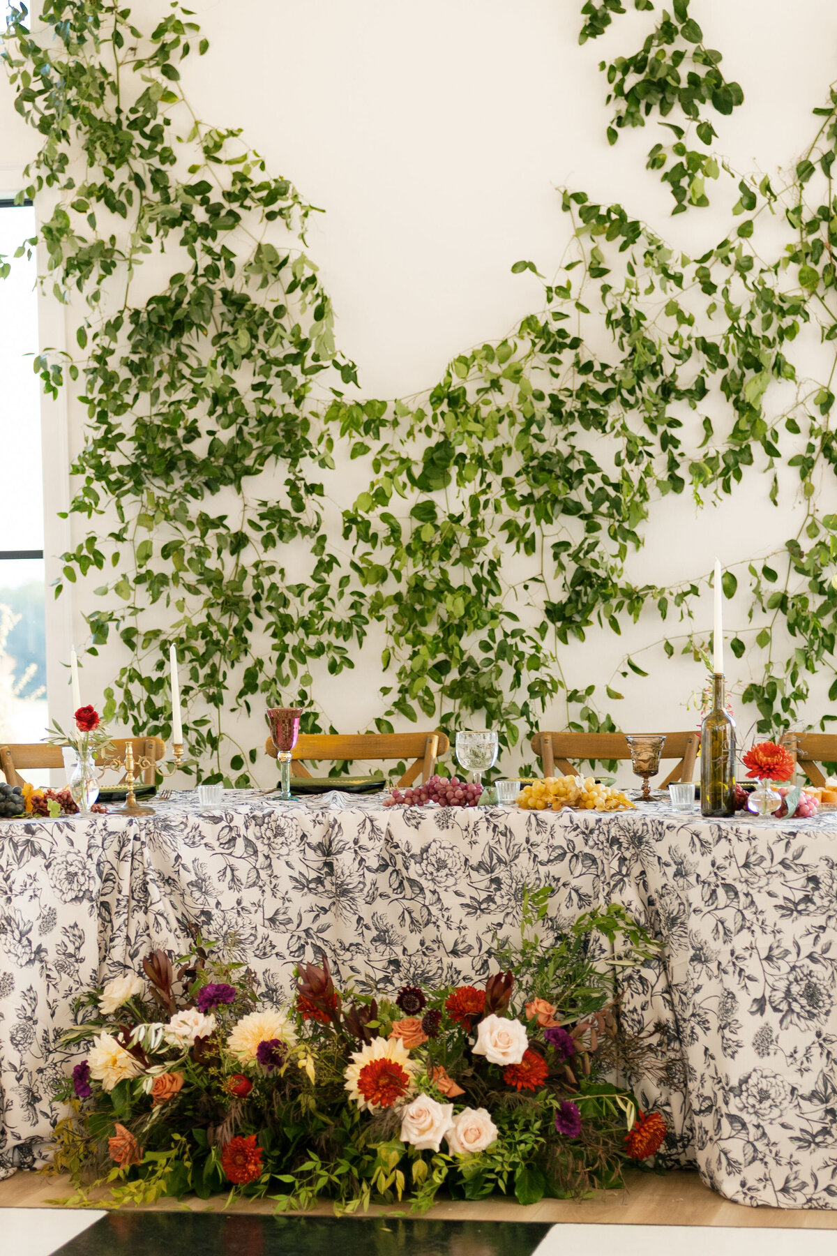 Wedding head table styled with black-and-white floral linens, abundant fruit accents, colorful glassware, and a lush floral ground arrangement, set against a dramatic greenery-covered wall for garden-inspired reception décor.
