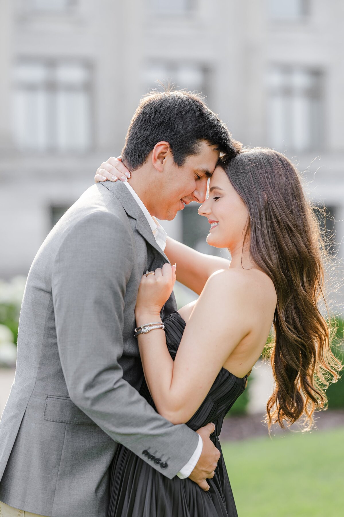 A newly engaged couple snuggles and holds each other in a lawn at sunset in a black gown and grey sports coat