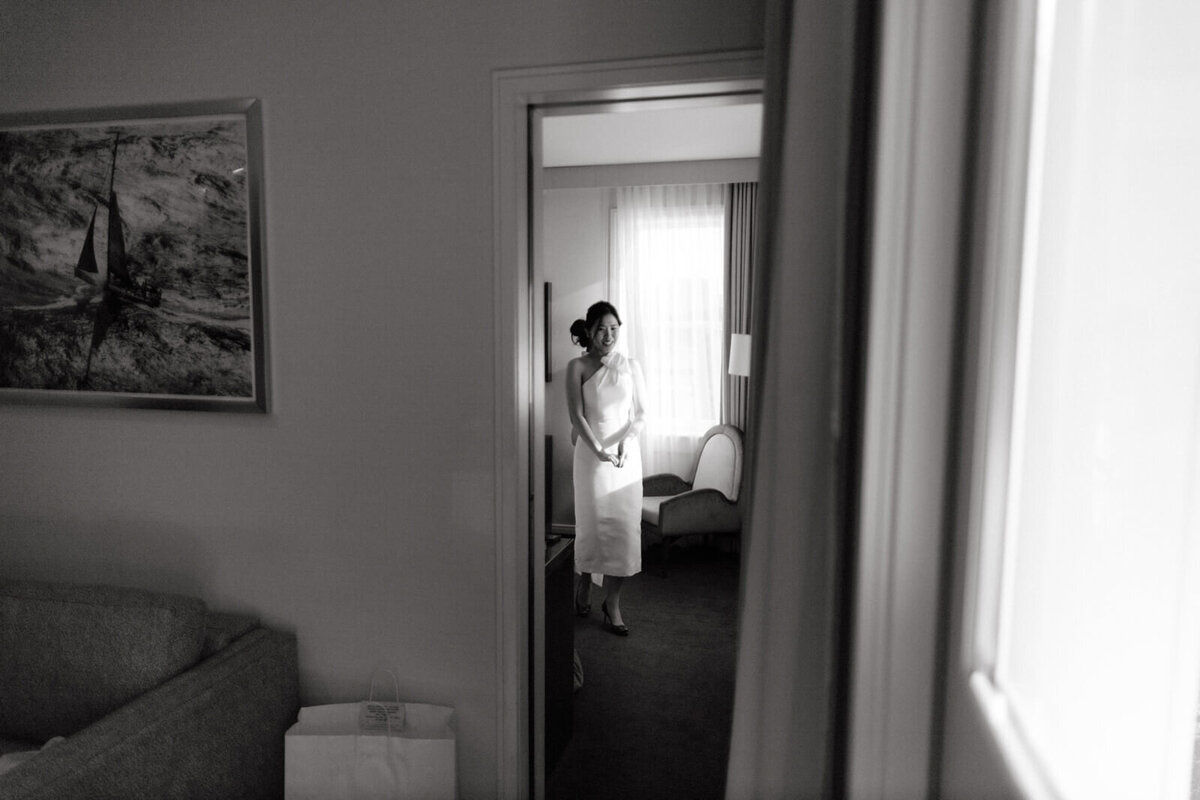 Black and white photo of a bride walking out of the room, with a couch and a painting outside of the room.