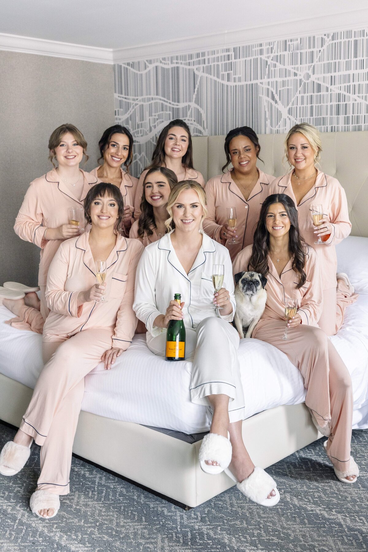 A group of nine women in matching pajamas sit and stand on a bed, holding champagne glasses. One woman sits in the center holding a champagne bottle. A pug is also on the bed. They appear to be celebrating and smiling.