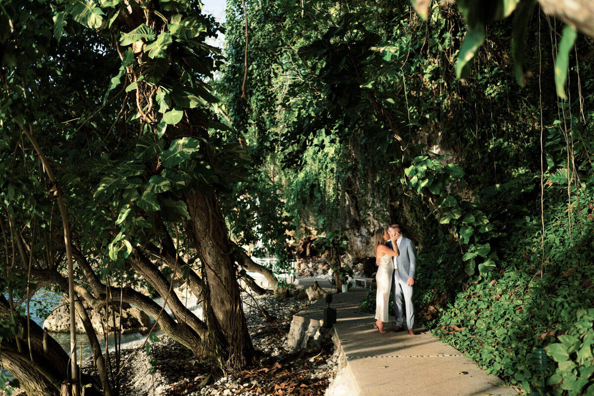 The engaged couple is kissing in a walking path with large trees around them, at Round Hill Hotel and Villas in Jamaica.