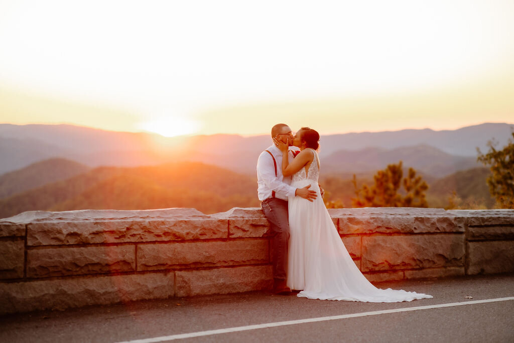Gatlinburg photographers captures sunset Foothills Parkway elopement with groom sitting on a stone wall while his bride holds his face and kisses him passionately with the Smoky Mountains in the distance