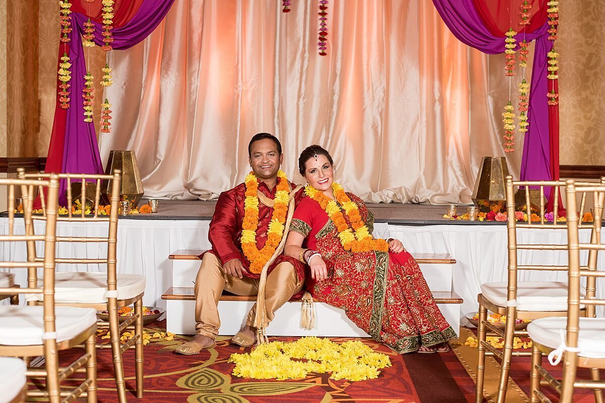 Indian bride and groom sitting under red and magenta mandap draped in strands of flowers wearing orange and yellow varmala garlands with flower petals on the floor and gold chivari chairs in the foreground