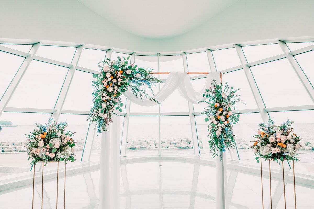 A beautiful wedding ceremony arbor with florals and white linens in front of a wall of windows