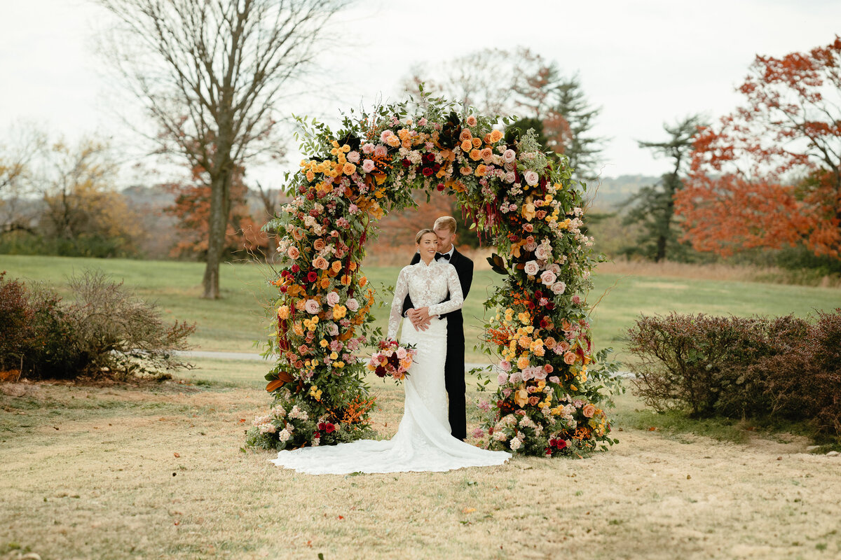 Bride and Groom standing underneath a floral arch covered in orange red and pink flowers