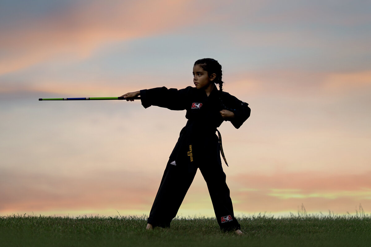 A young girl performs karate at sunset in a black  outfit in a park with a stick straight out