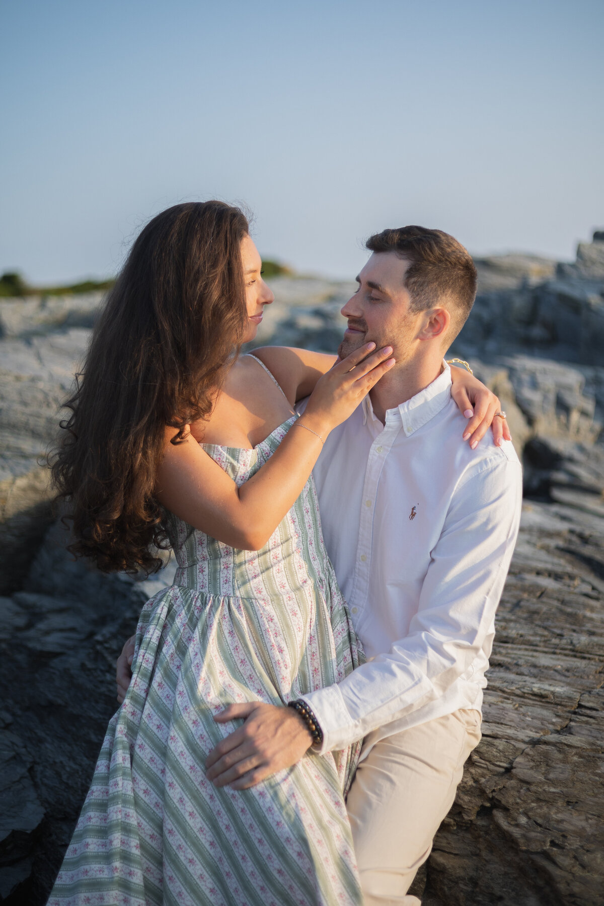 Collins Beach Newport RI | Kelsey Sheehan Photography Timeless Rhode Island Weddings | A couple sits on rocky terrain by the sea, smiling and embracing warmly. The woman in a floral dress gently touches the man's face, conveying affection.