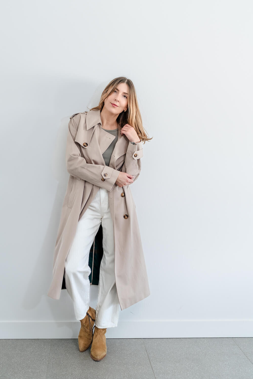Stylish woman in beige trench coat, white pants, and suede boots leaning casually against white wall
