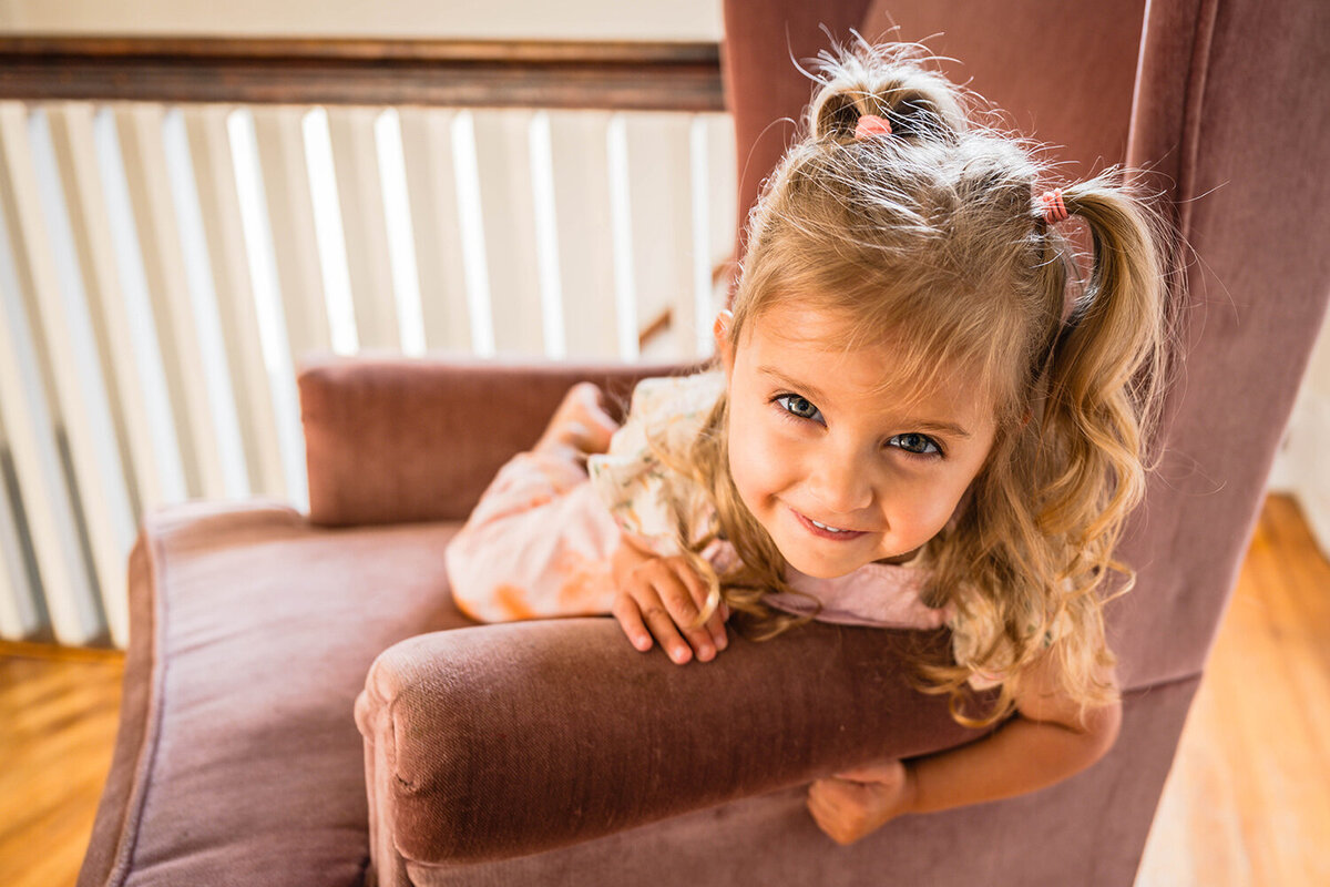 A toddler hangs over the side of a chair, smiling widely, for a portrait.