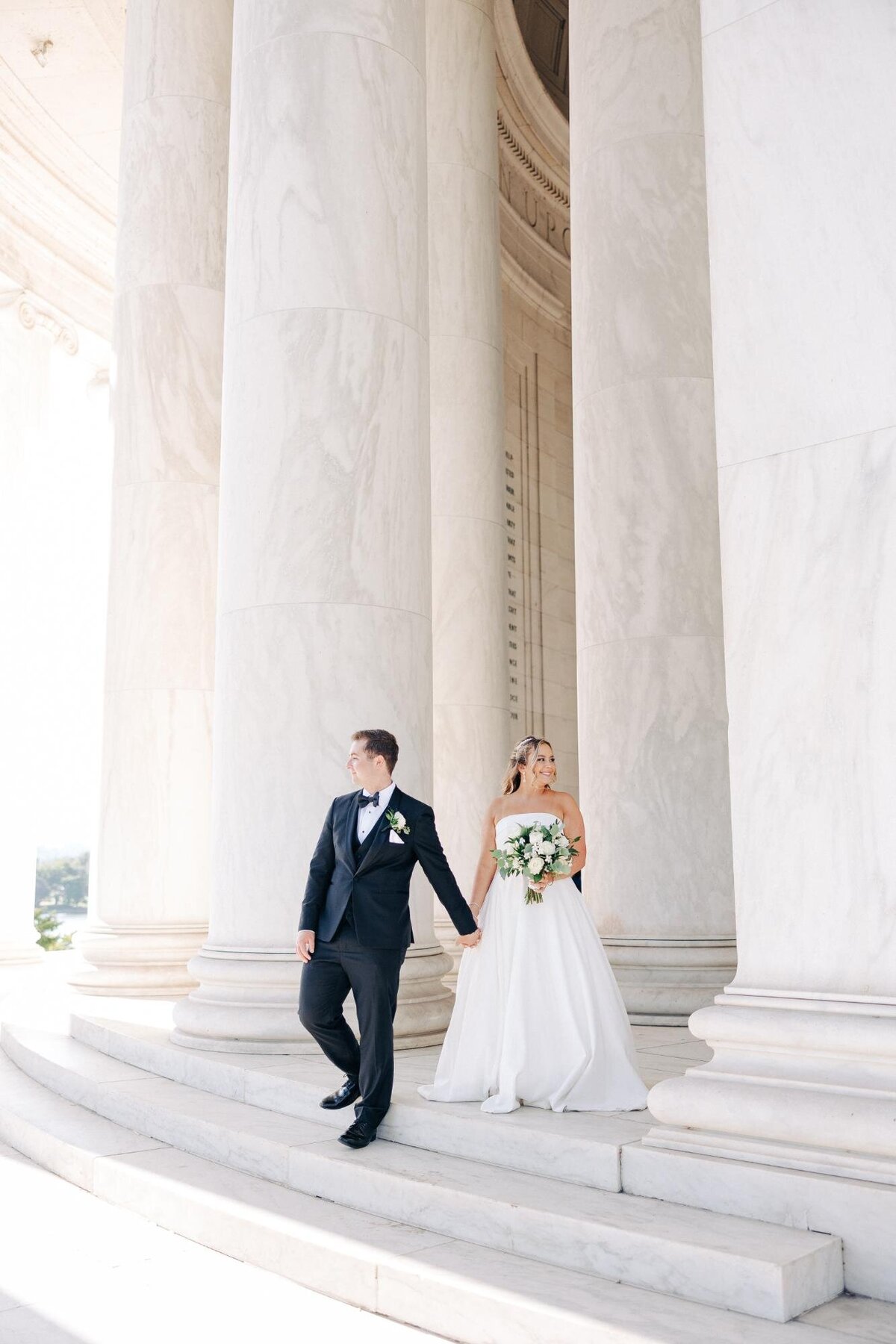 A wedding couple holding hands beside large pillars, with the groom looking at the bride as they both stand in natural light.
