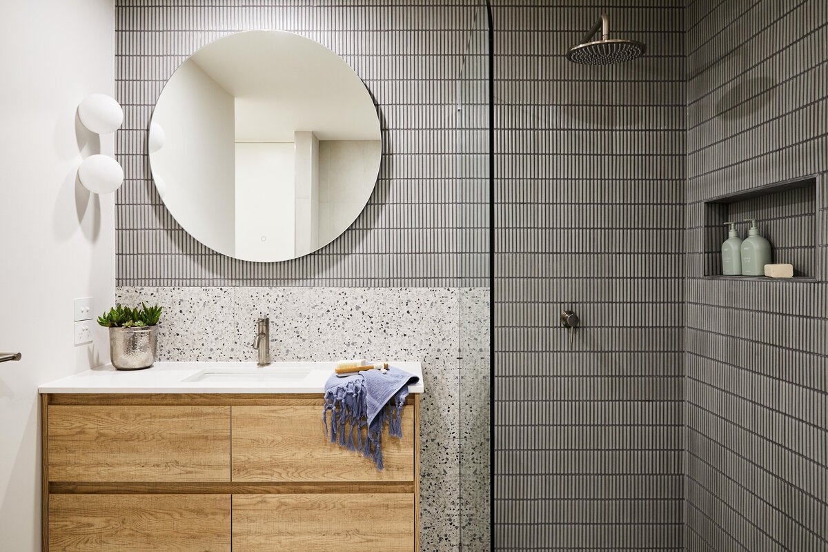 Wider view of the Elwood bathroom showing a circular mirror above a timber vanity and a walk‑in shower lined with vertical grey kit‑kat tiles and a recessed shelf.