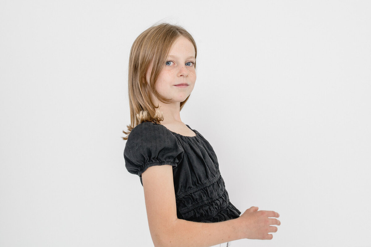 A young blonde girl with short hair wearing a black dress in front of a white background.