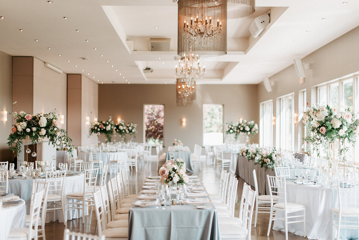 A combination of long harvest tables and round tables draped with dusty blue linen  and a combination of tall and low centrepieces in dusty blush fill the modern reception space at Le Belevedere for a wedding in Wakefield Quebec