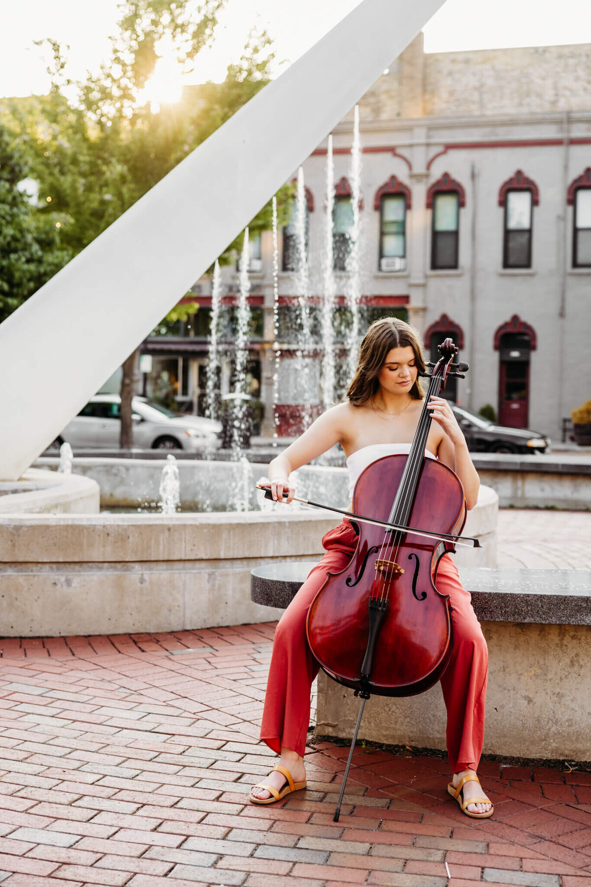 stunning teenage girl in a white top and red pants playing her cello in front of a fountain in downtown Green Bay. 