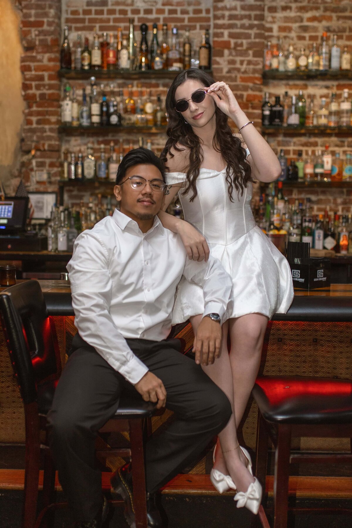  stylish couple poses confidently inside a bar with an exposed brick wall and shelves of liquor in Jacksonville, Florida. The bride-to-be wears an off-the-shoulder white satin mini dress and sunglasses, while her fiancé leans casually on the bar stool in a crisp white shirt and dark pants. Their bold, editorial look gives off a cool, modern vibe. Captured by Heartful Films, a Jacksonville engagement photographer and videographer known for creative, personality-driven sessions that break the mold.