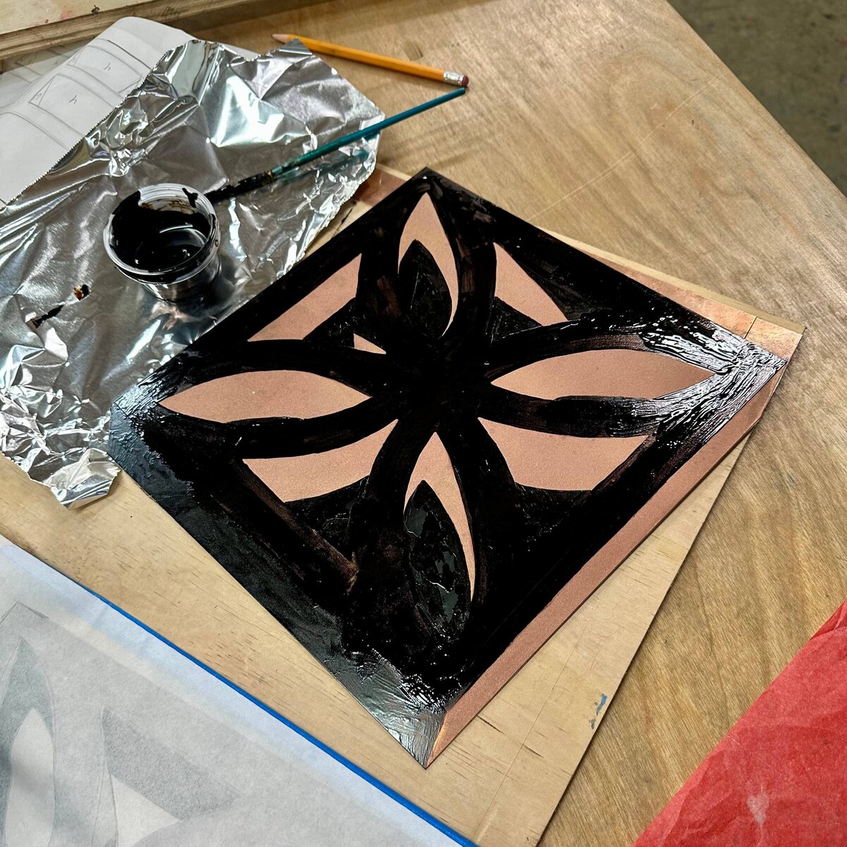 A copper plate with hand-painted asphaltum resist in bold floral shapes rests on a wood surface next to a brush and foil palette.