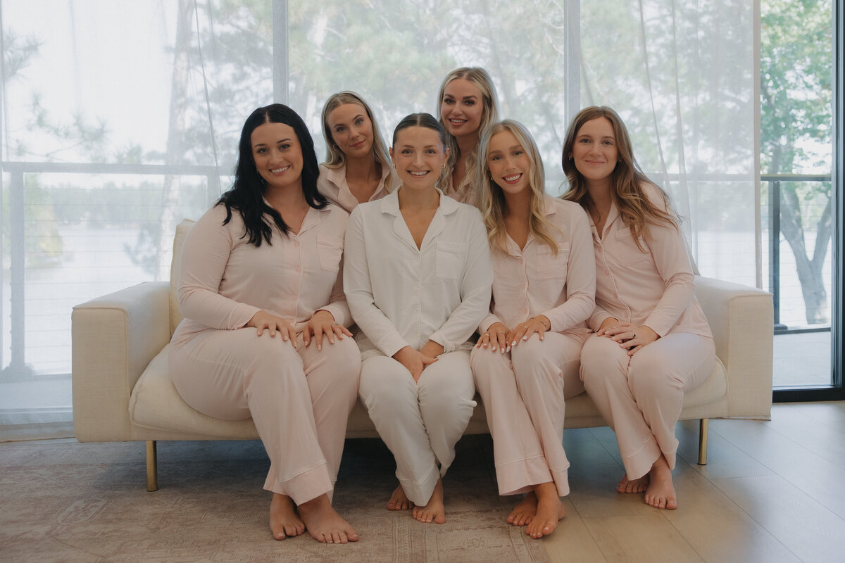 smile sitting on the couch. There are 4 girls sitting on the main part of the couch and two girls up on the back rest of the couch crouching down. There is a wall of windows behind the couch covered in a slightly see through curtain.